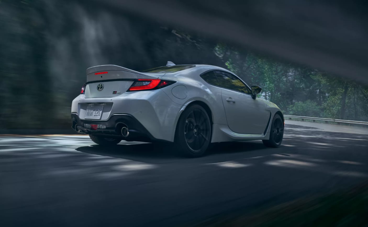 A white Toyota GR86 is shown from the rear as it speeds past the viewer with a blurry, dark forest scene behind