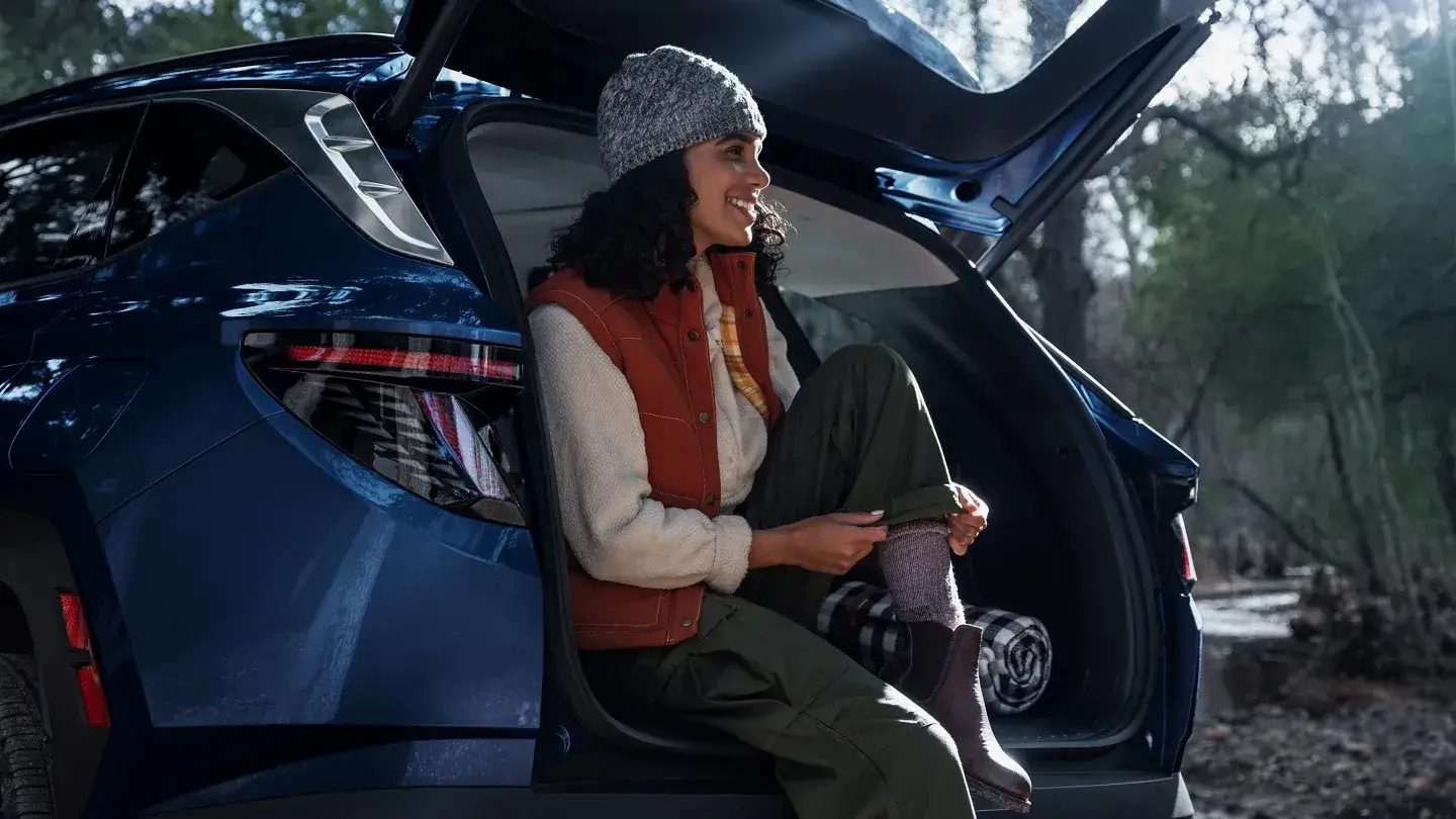Woman sitting in the open cargo area of a blue Hyundai Tucson Limited in a forest, rear three-quarter view