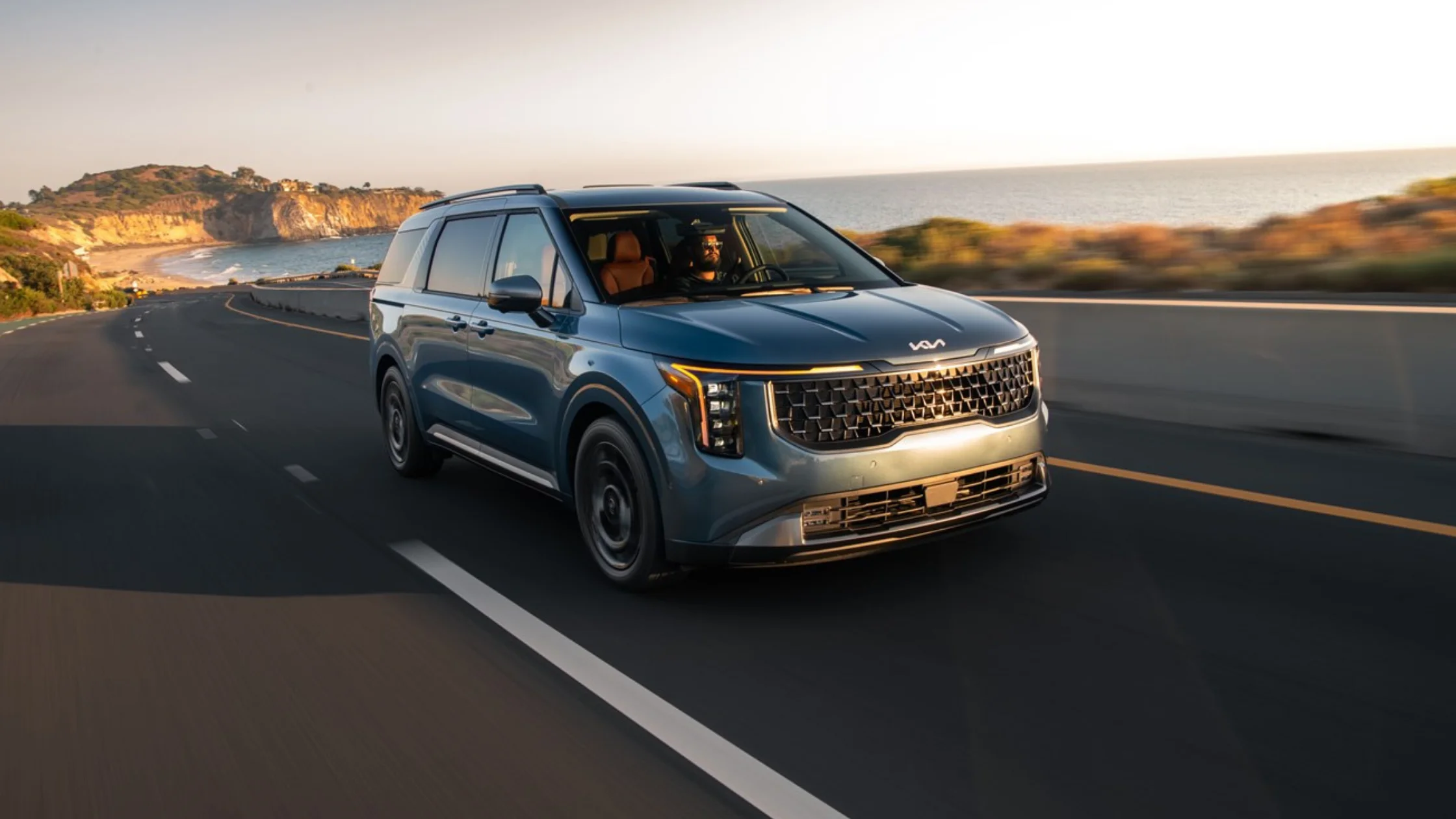 2026 Kia Carnival minivan driving along a coastal highway at sunset with ocean cliffs in background