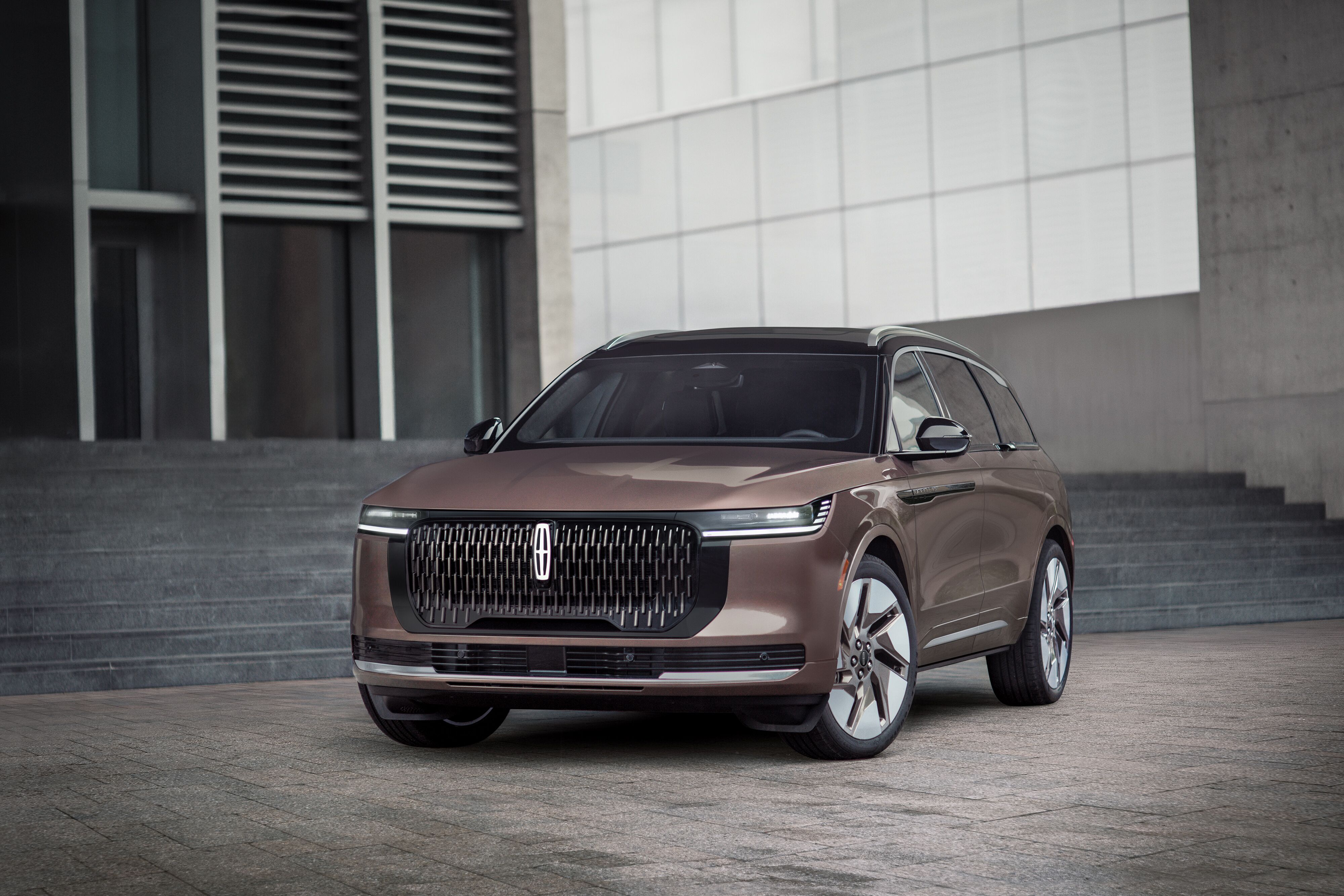Front three-quarter view of a metallic copper 2027 Lincoln Nautilus luxury midsize SUV parked outside modern architecture.