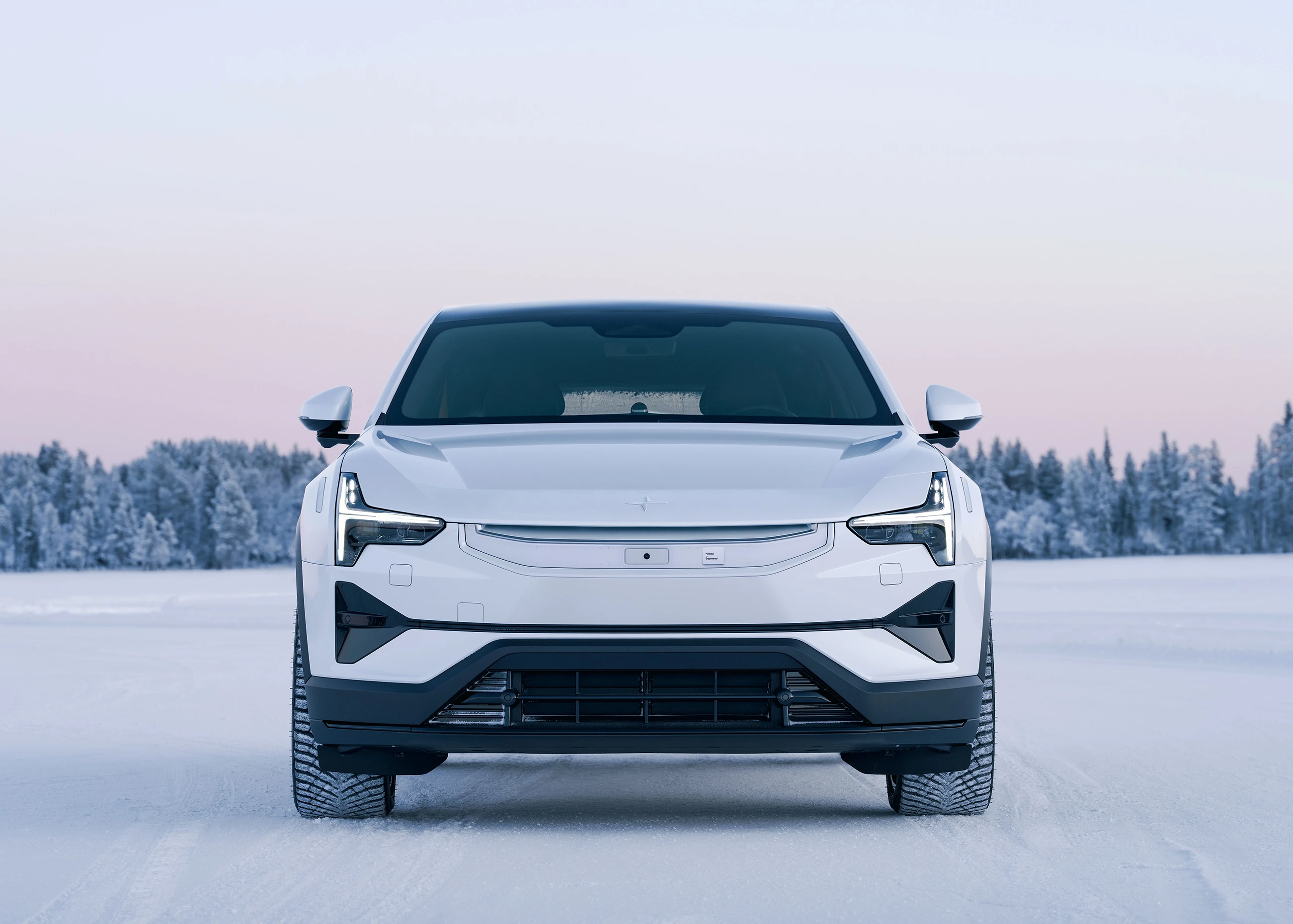 Head-on view of a Polestar 3 parked on a frozen lake with a snow-covered tree line in the distance