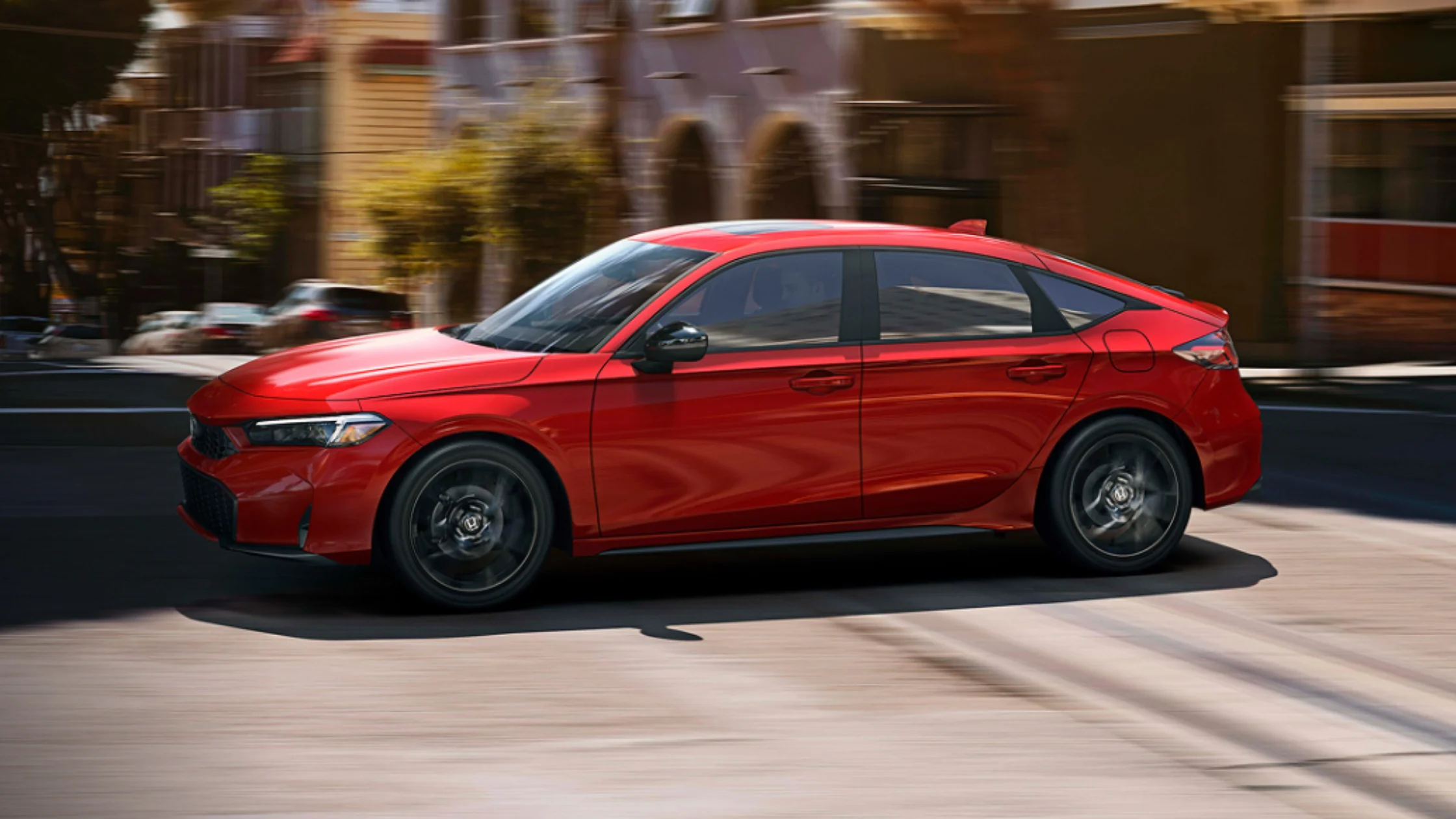 2026 Honda Civic Hatchback in red driving through city street, showcasing sporty silhouette and alloy wheels.