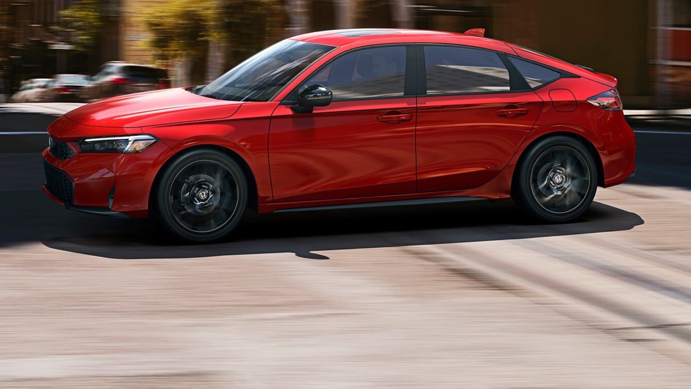 2026 Honda Civic Hatchback Hybrid in red driving through the city with black wheels and sporty look.