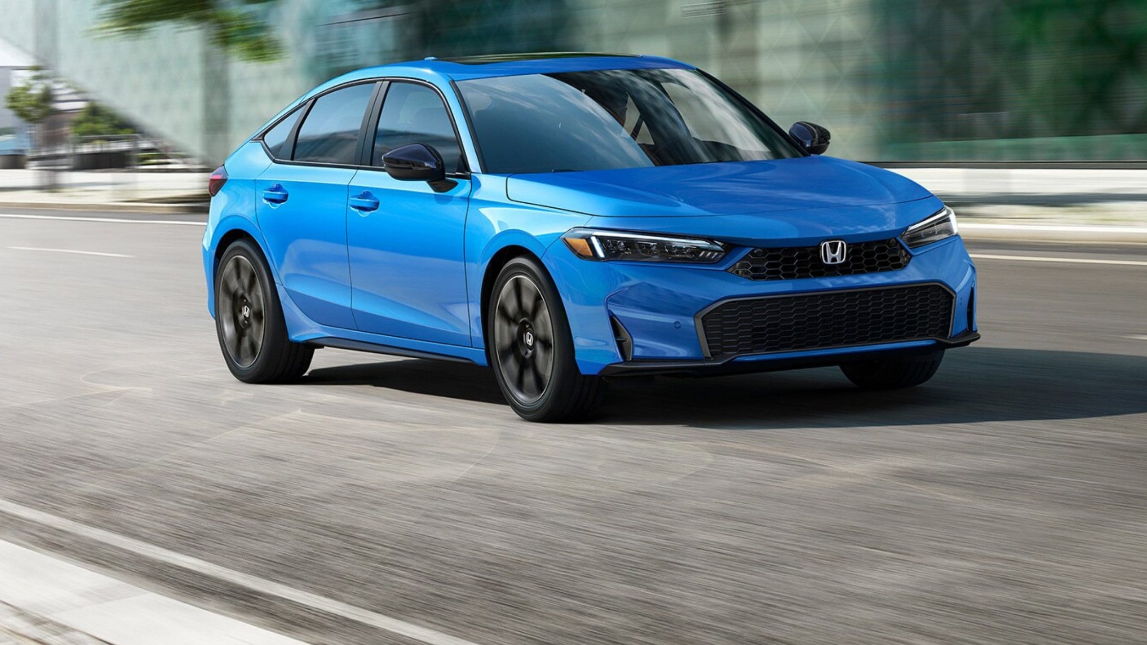 2026 Honda Civic Hatchback in blue driving on a city road with black wheels and sleek front styling.