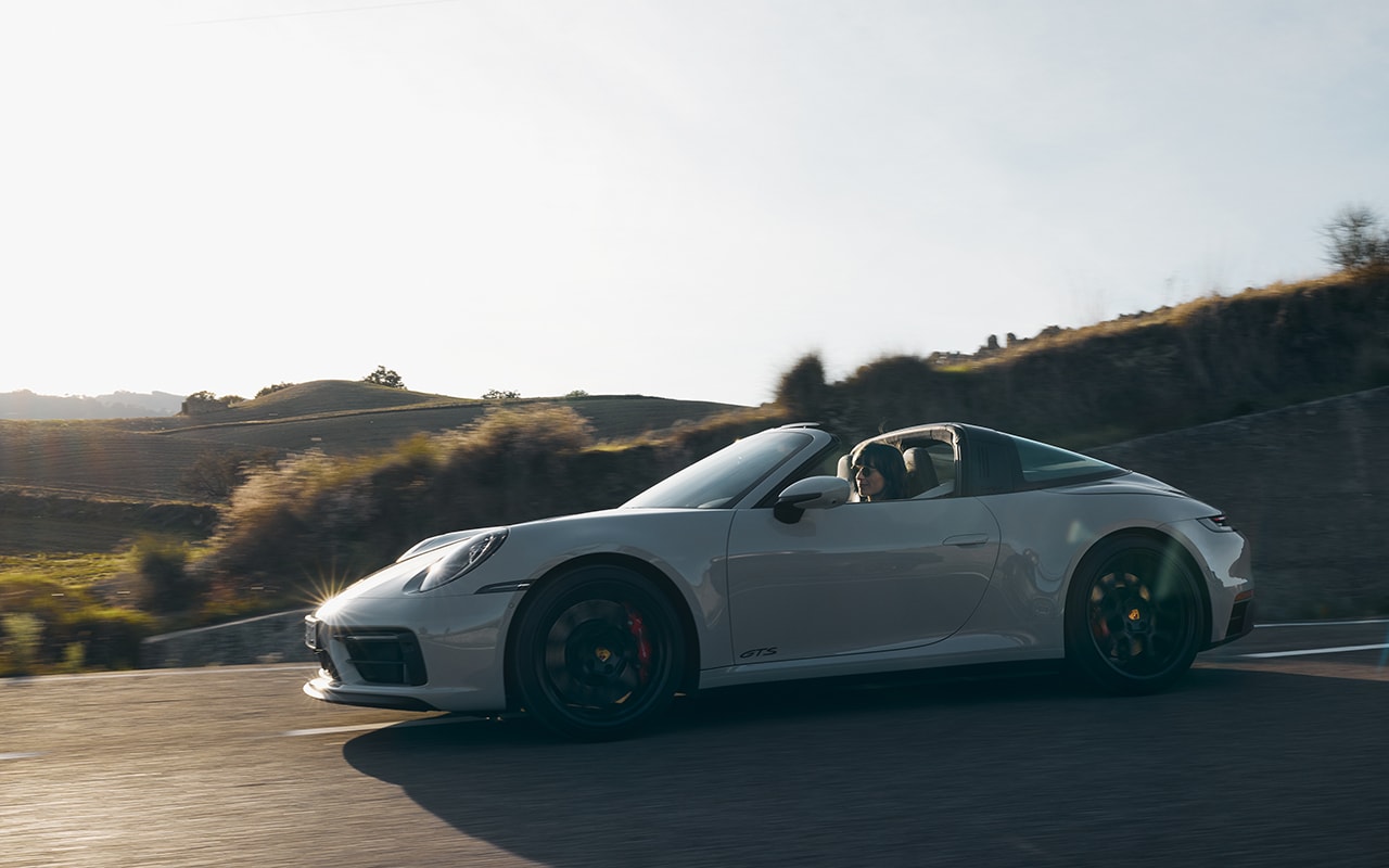 woman driving a white Porsche 911 fast next to dry hills