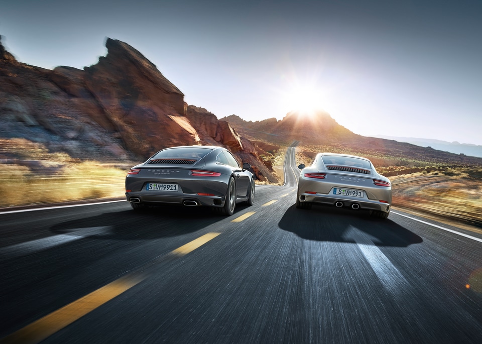 two Porsche 911 Carrera coupes driving along a desert highway, side-by-side