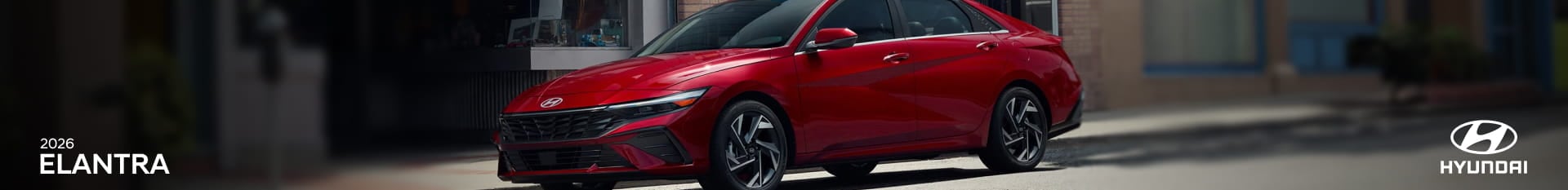 A sleek red 2026 Hyundai Elantra is parked on a sunlit street in front of storefronts.
