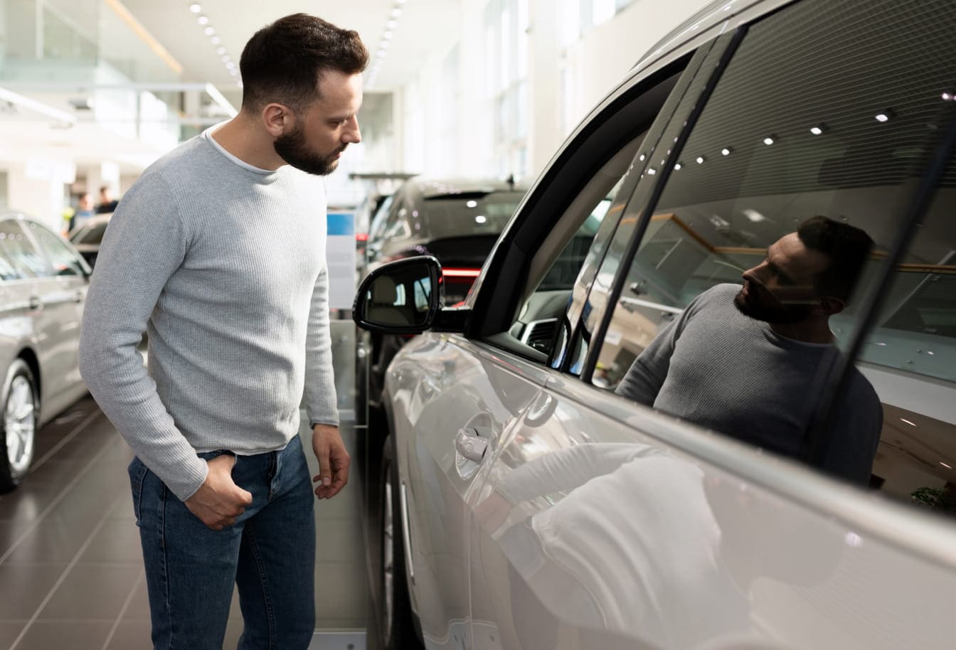 A man examines a car's exterior in a showroom, reflecting on the glossy surface. The setting is bright and modern.