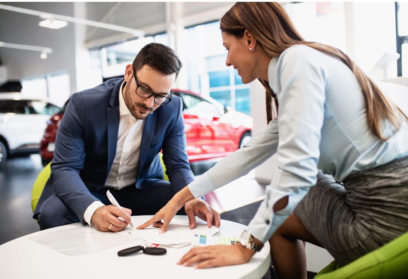 A man and woman discuss financing in a car dealership, sitting at a table. The woman points at the document, while the man writes, appearing focused.