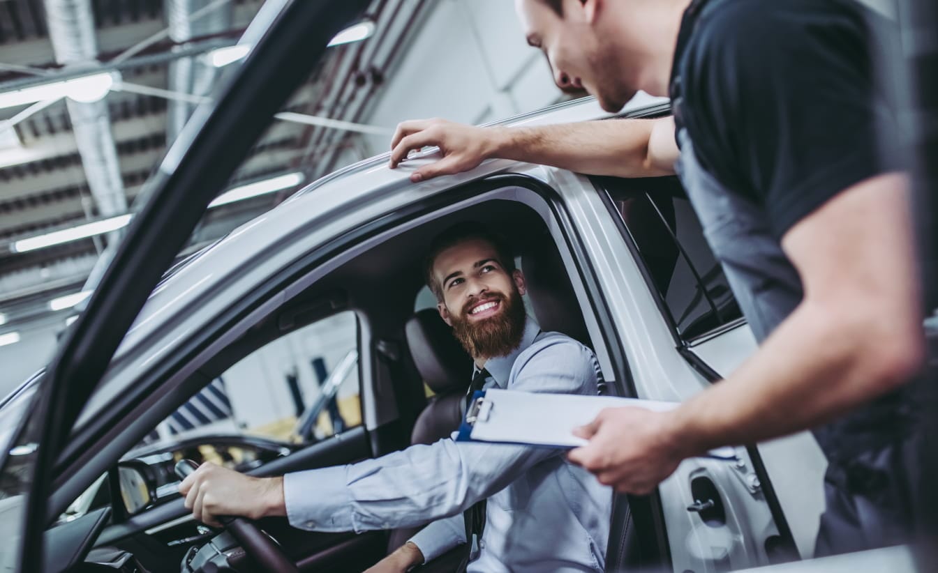Man in a car smiles at a mechanic holding a clipboard inside a garage. The interaction appears positive, reflecting a friendly and professional service.