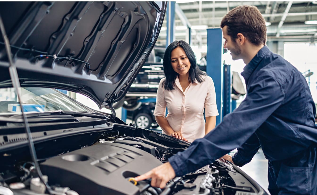 Mechanic in blue coveralls explains car engine details to a smiling woman in a white shirt in a garage. The scene conveys professionalism and trust.