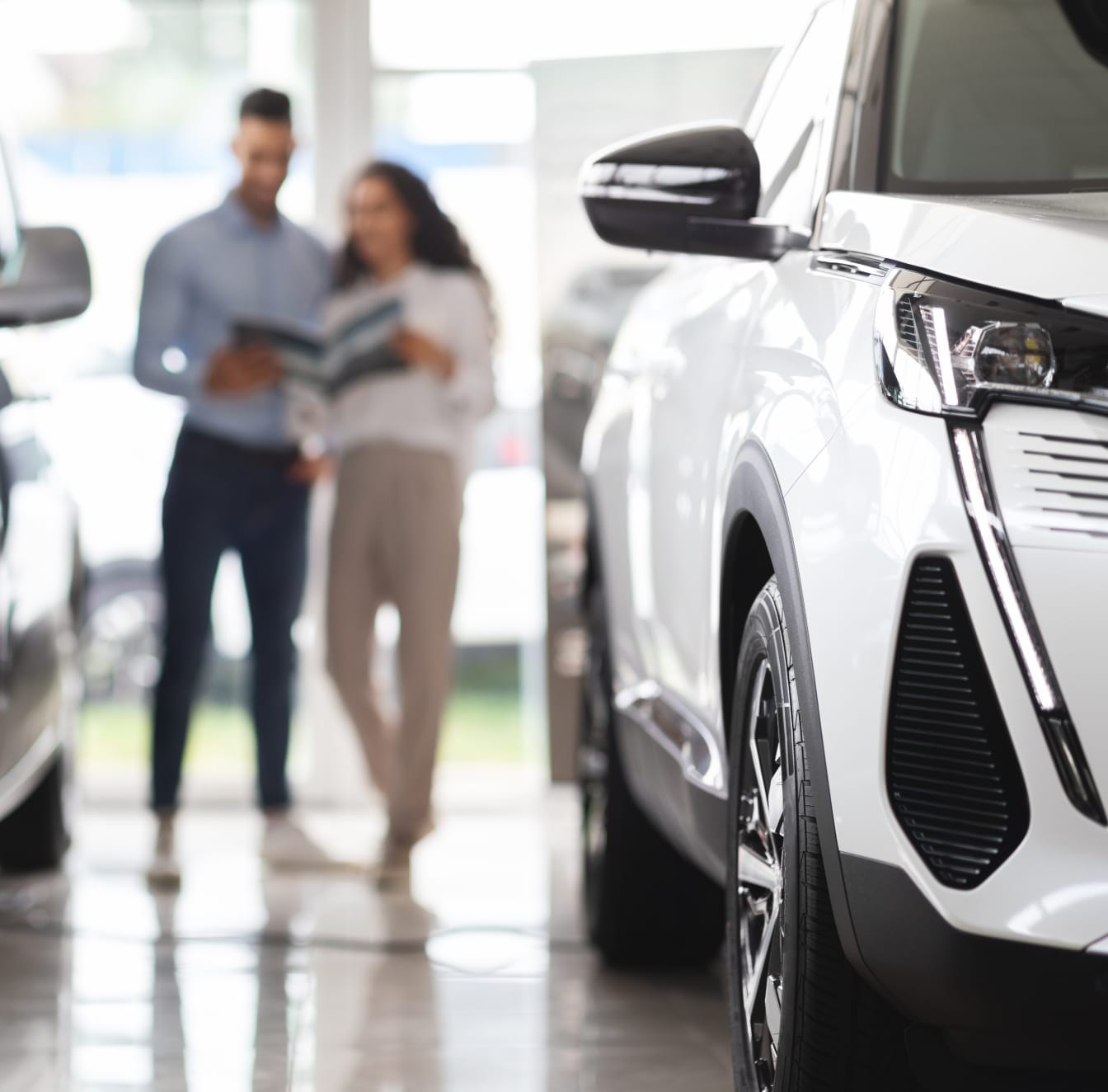 Close-up of a white car's front in a showroom, with a blurred couple in the background holding a brochure, suggesting interest in purchasing.