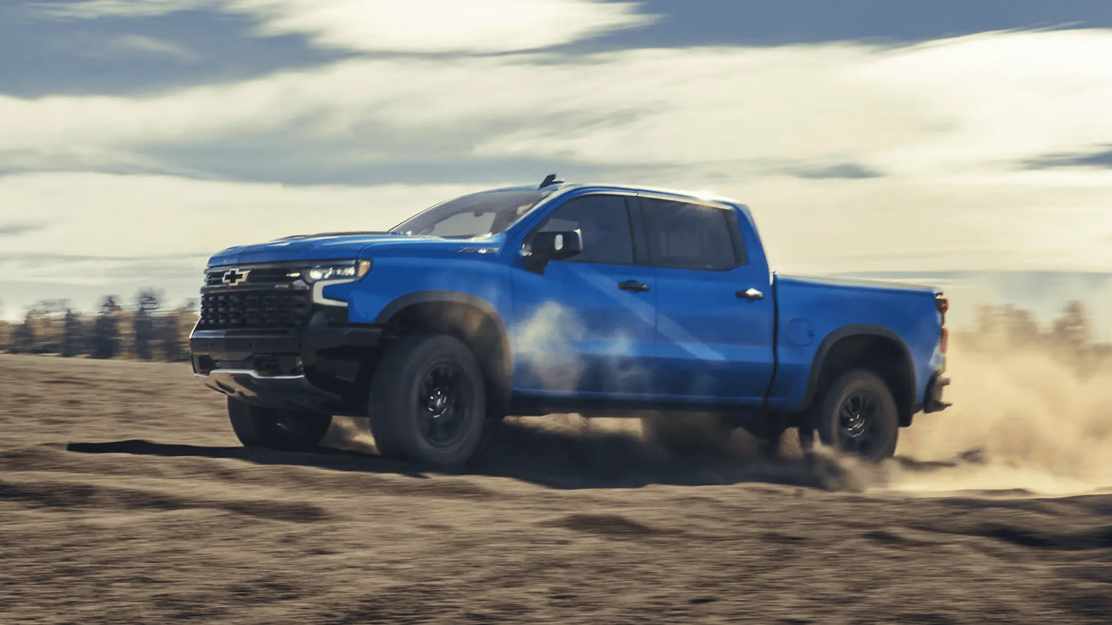 A blue 2025 Chevrolet Silverado pickup truck kicking up dust on a rugged off-road terrain.