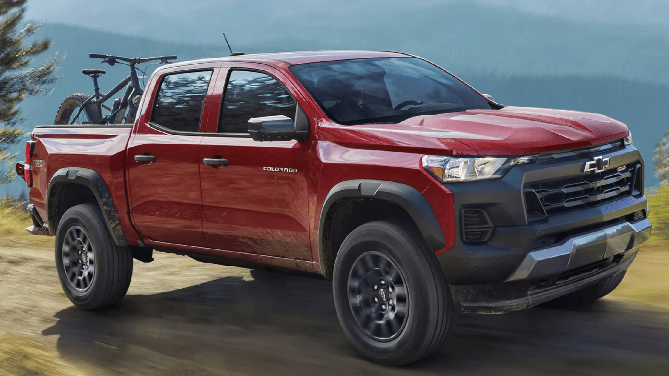 A red 2025 Chevrolet Colorado pickup carrying mountain bikes in the bed, driving on a dirt path.