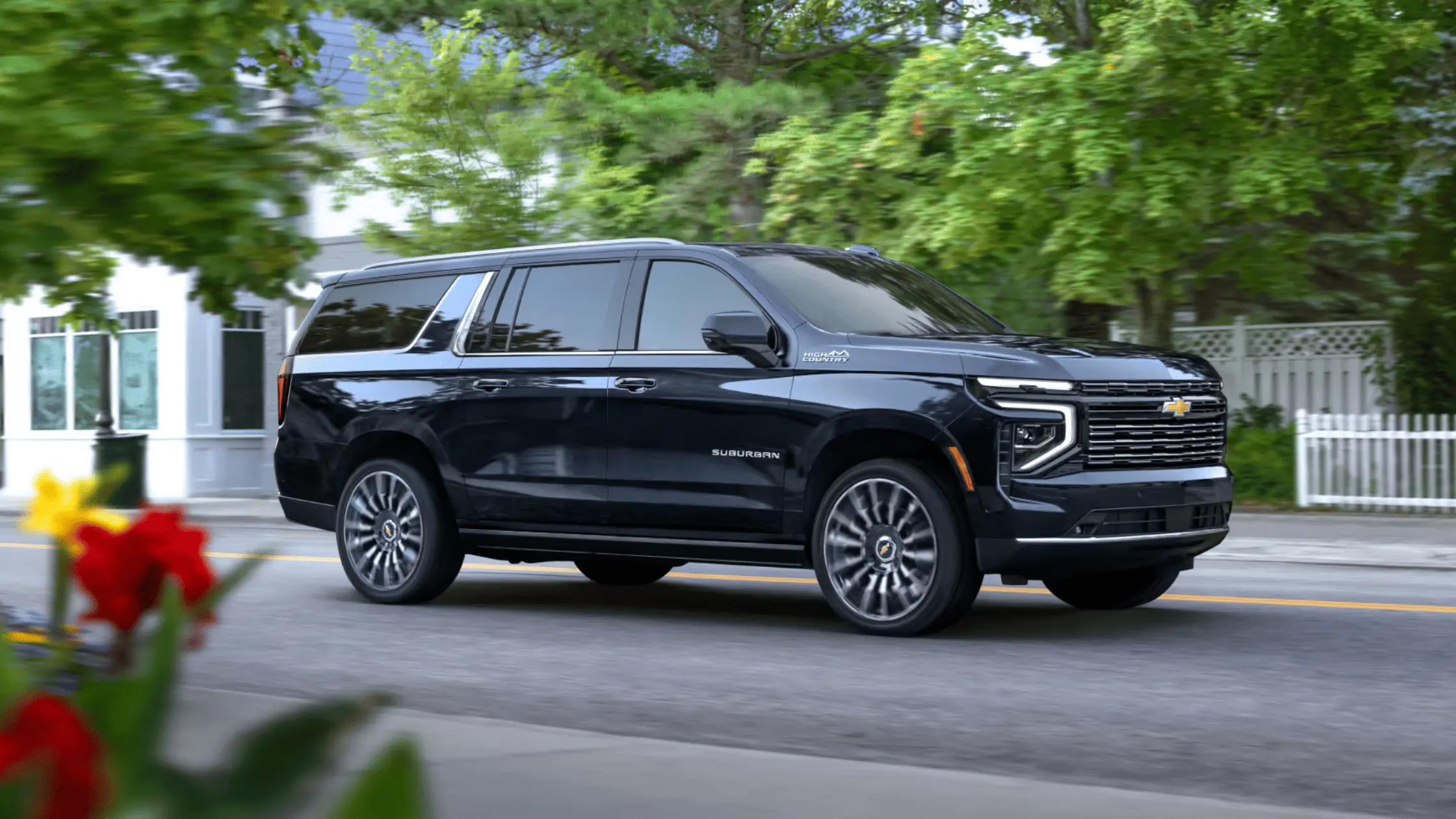 A black 2025 Chevrolet Suburban High Country driving through a residential neighborhood lined with trees.