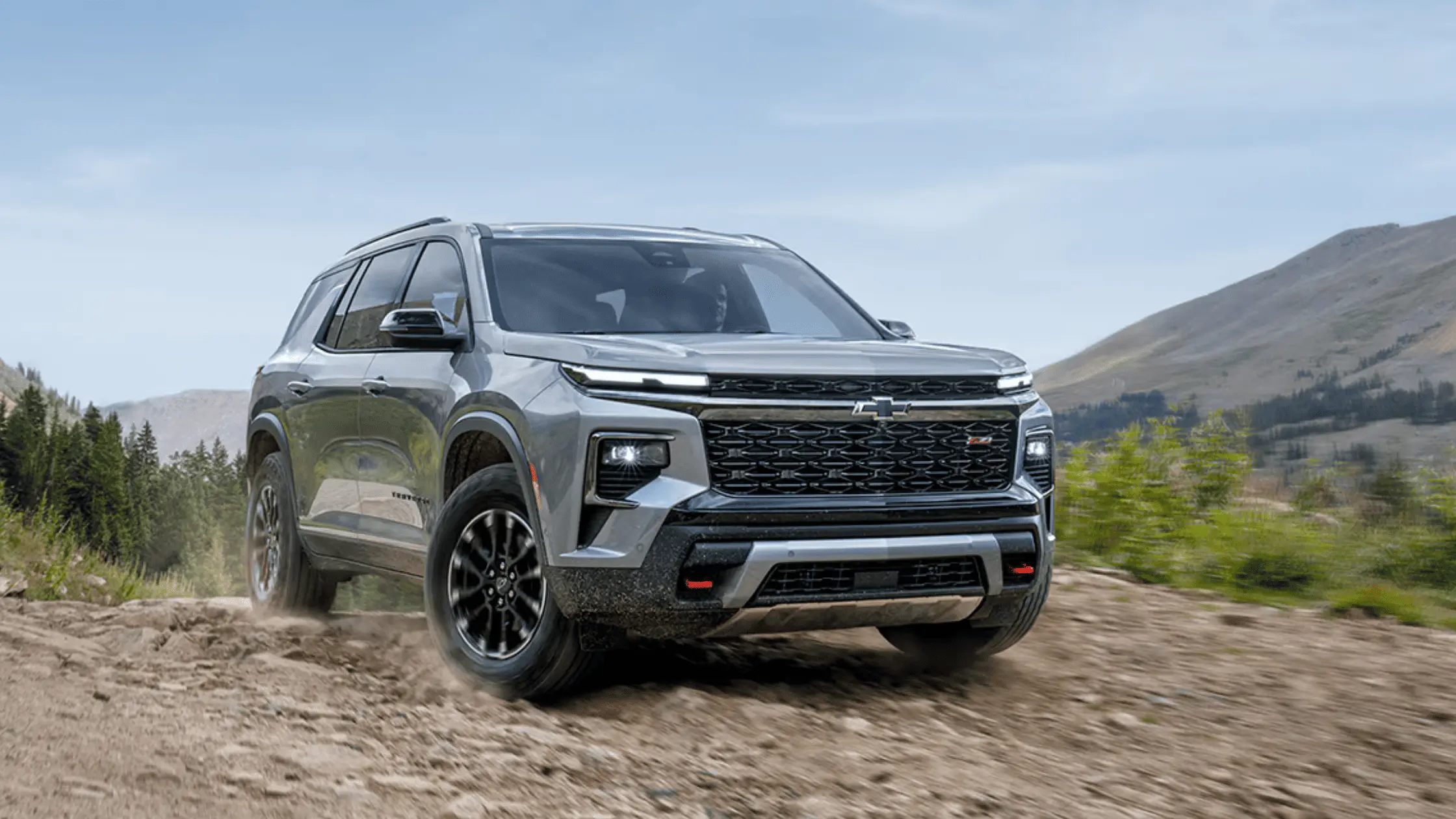 A silver 2025 Chevrolet Traverse SUV driving on a rocky off-road trail with mountains in the background.