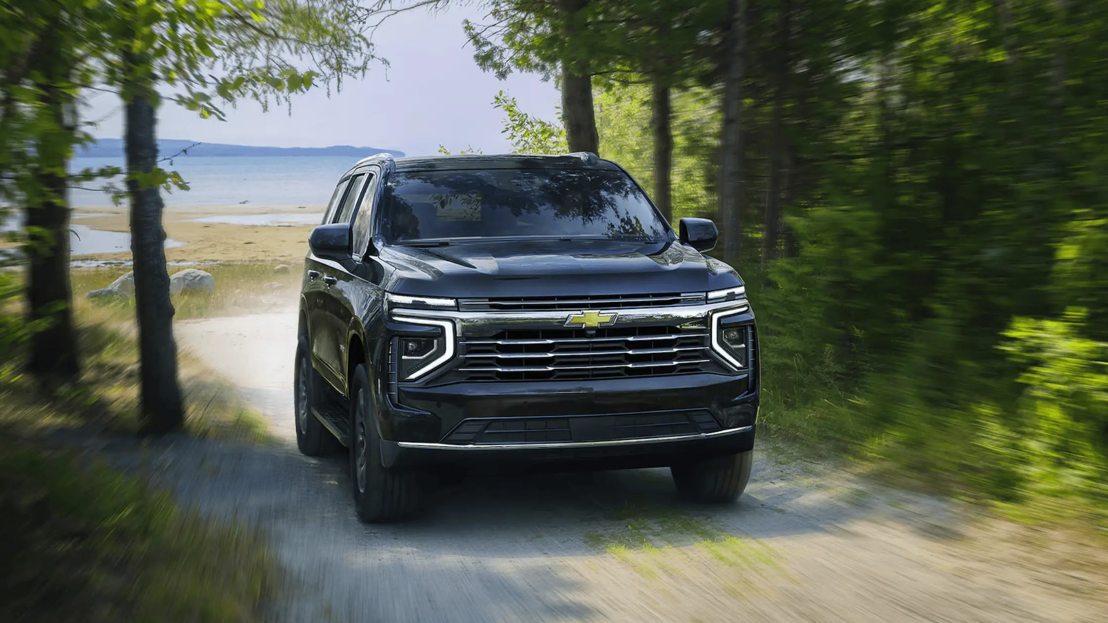 A black 2025 Chevrolet Tahoe driving on a forest trail near a lake or beachfront in bright daylight.
