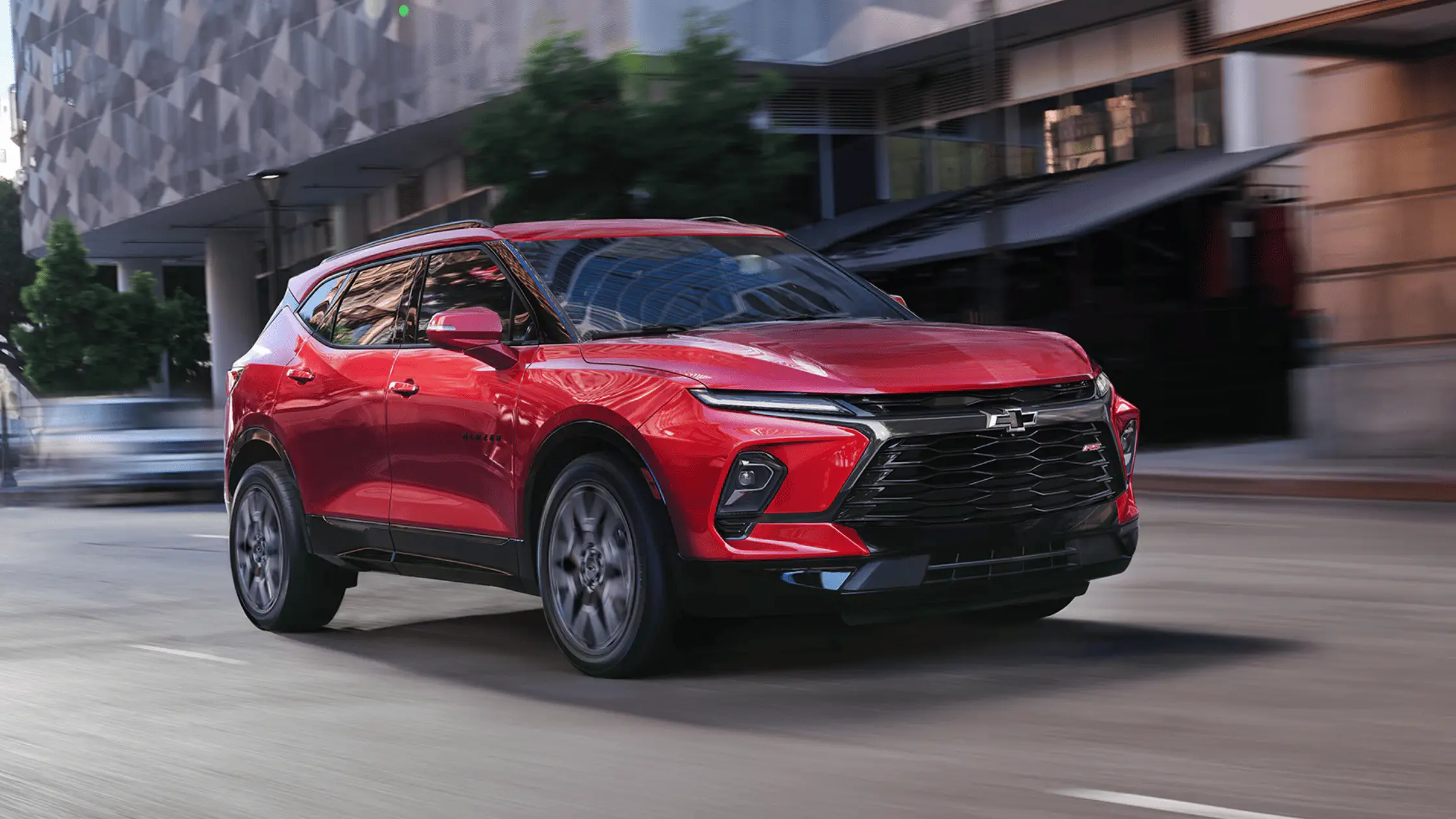 A red 2025 Chevrolet Blazer RS driving through a downtown city street with modern architecture.