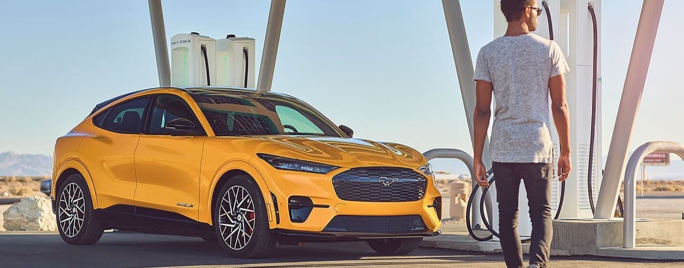 A person walking by a yellow 2024 Ford Mustang Mach-E GT plugged into a charging station.