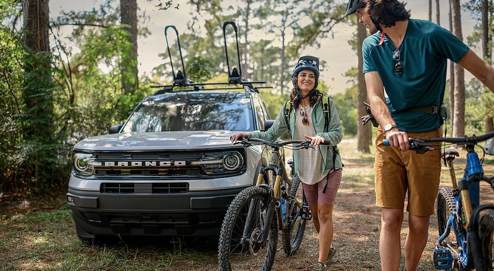 Two people walking their bikes past a white 2023 Ford Bronco Sport parked by a trailhead.