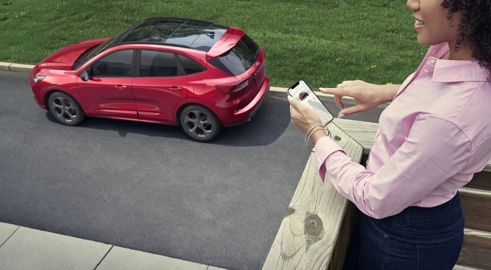 A woman on a balcony checking the status of a red 2023 Ford Escape using a smartphone app.