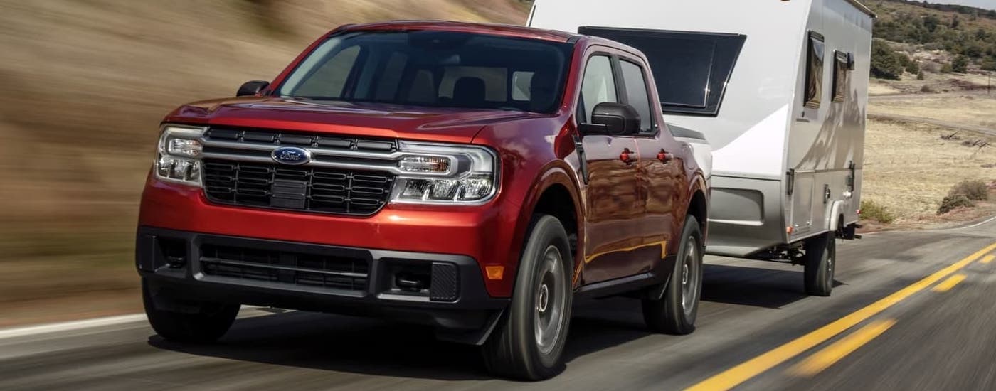A red 2024 Ford Maverick Ecoboost Lariat is shown towing a trailer on an open road.