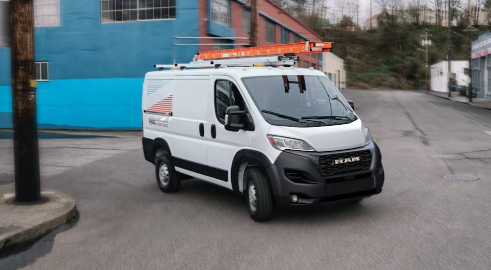 A white 2024 Ram ProMaster Cargo Van with ladders on the roof driving around a corner.