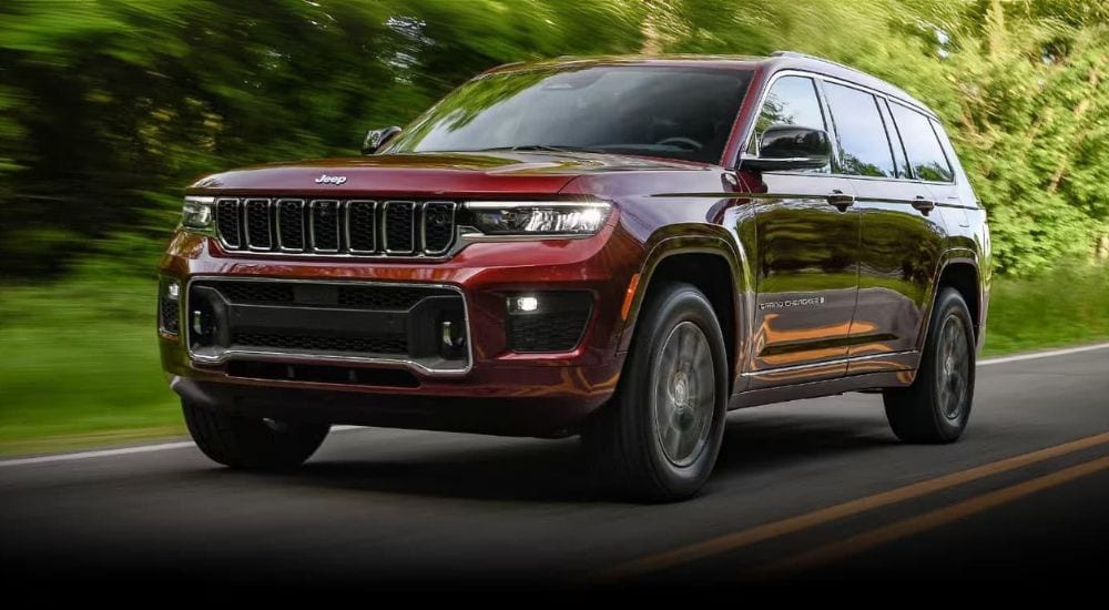 A red 2024 Jeep Grand Cherokee is shown driving on a tree-lined road after leaving a Jeep dealer.