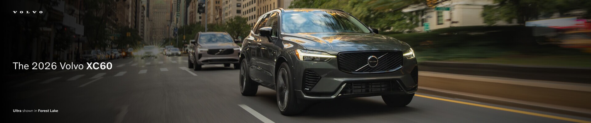 A front view action shot of The 2026 Volvo XC60 Ultra in Forest Lake green driving down a city street.