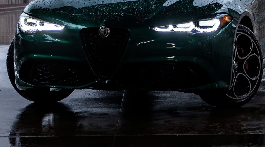Front view of a green 2026 Alfa Romeo Giulia with glowing LED headlights and rain droplets on wet asphalt for autumn wet weather driving near Ramsey, NJ
