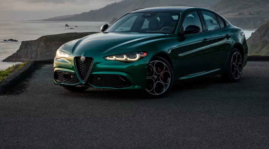 Front three-quarter view of a metallic green 2026 Alfa Romeo Giulia with illuminated LED headlights glowing on an asphalt overlook against a dramatic coastal cliffs backdrop. The detailed shot highlights the signature Scudetto triangular grille, dark 5-hole phone-dial wheels, red brake calipers, and Veloce trim badge, demonstrating 2026 Alfa Romeo Giulia performance and Q4 AWD capability for Bergen County drivers on Route 17.