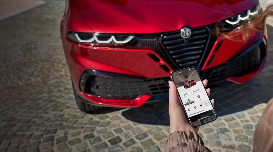 Driver holding a smartphone displaying the myAlfa Romeo app to monitor battery level and EV charging schedules in front of a red 2026 Alfa Romeo Tonale PHEV in Bergen County, NJ.