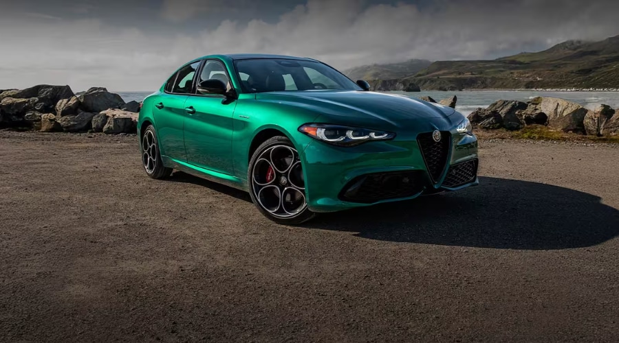 A metallic green 2026 Alfa Romeo Giulia luxury sports sedan parked on a gravel overlook against a scenic coastal backdrop. The front three-quarter shot highlights the signature triangular Scudetto grille, dark 5-hole alloy wheels with red brake calipers, and aerodynamic styling, showcasing 2026 Alfa Romeo Giulia capability and Q4 AWD performance for Bergen County commuters.