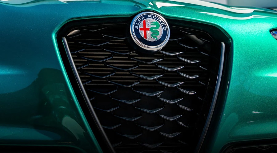 Extreme macro close-up of the iconic triangular Scudetto front grille and colorful roundel emblem on a metallic green 2026 Alfa Romeo Giulia luxury sports sedan. The detailed shot highlights the black V-mesh grille pattern, red cross, and Biscione serpent badge against vibrant Verde green paint, showcasing authentic Italian craftsmanship at Ramsey Alfa Romeo in Bergen County, NJ.
