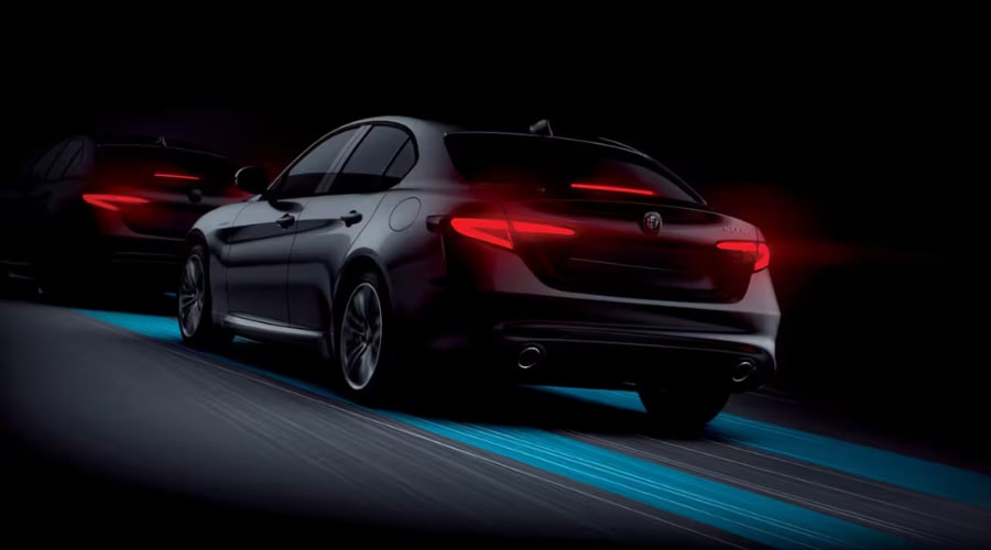 Rear three-quarter action shot of a dark metallic 2026 Alfa Romeo Giulia driving at night with glowing red LED taillights, dual exhaust ports, and blue lane-tracing graphic lines beneath the tires, illustrating Active Driver-Assist safety features and Q4 AWD stability for night commutes along I-287 and Route 17 in Bergen County, NJ.