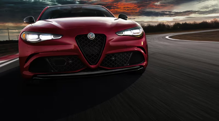 Low-angle front action shot of a deep crimson red 2026 Alfa Romeo Giulia performance sports sedan driving dynamically along a curved asphalt road at dusk. The dramatic angle highlights glowing LED Matrix headlights, iconic triangular Scudetto V-shield grille, carbon fiber front lip spoiler, and sculpted hood, showcasing 2026 Alfa Romeo Giulia capability and handling for Bergen County commuters in Ramsey and Mahwah, NJ.