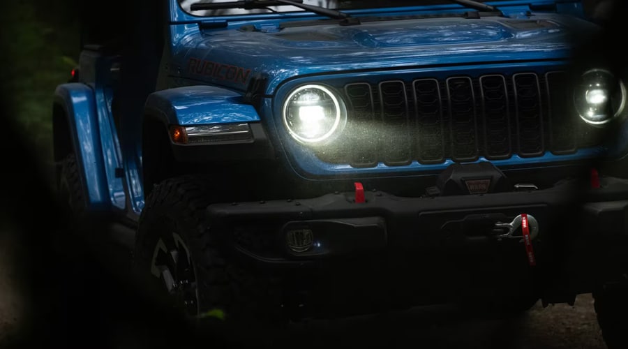 Close up front view of a blue 2026 Jeep Wrangler Rubicon showing glowing circular LED headlights, the iconic black seven slot grille, and a rugged steel bumper with an integrated winch on a dark wooded trail in Ramsey New Jersey