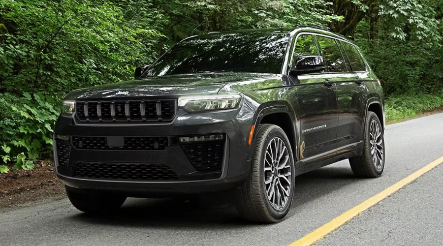 Dark metallic charcoal 2026 Jeep Grand Cherokee 4x4 SUV parked along a paved forest road surrounded by dense trees, illustrating Quadra-Trac drive maintenance for back-road commutes near Rockland and Bergen County, NJ.