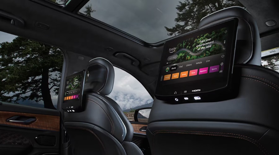 Rear seat interior perspective of a 2026 Jeep Grand Cherokee showing dual headrest-mounted Fire TV entertainment screens, black leather seats, and panoramic sunroof, highlighting cabin air filter care for comfortable family summer road trips in Bergen County, NJ.