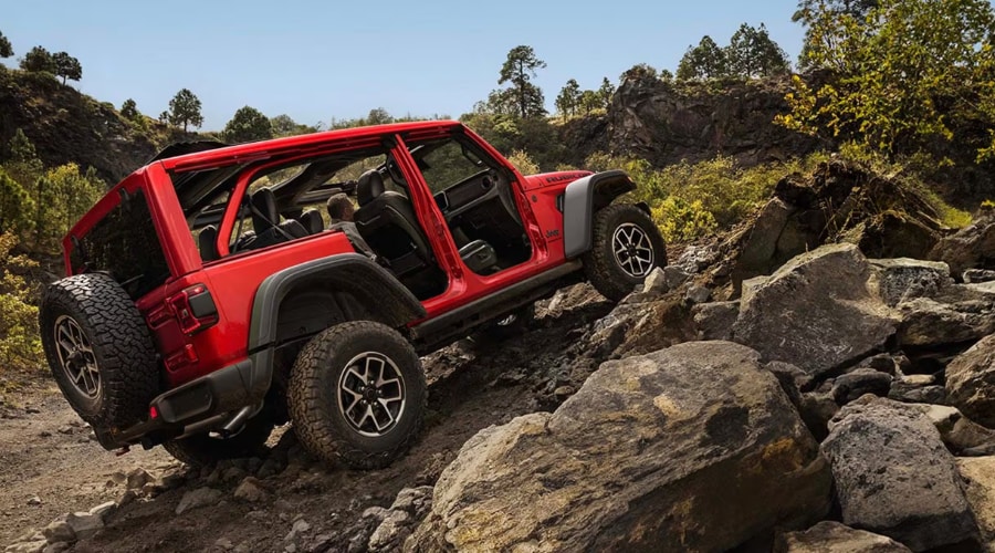 Rear three-quarter view of a red 2026 Jeep Wrangler Rubicon with its doors removed and top open, articulating its suspension while climbing up a steep incline over large, sunlit rocky boulders on an off-road trail near Ramsey NJ