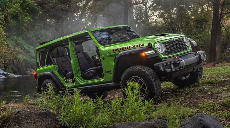 Three-quarter front view of a bright green 2026 Jeep Wrangler Rubicon with all doors removed, parked on a grassy, dirt path next to a flowing forest stream on a sun-dappled afternoon near Ramsey NJ