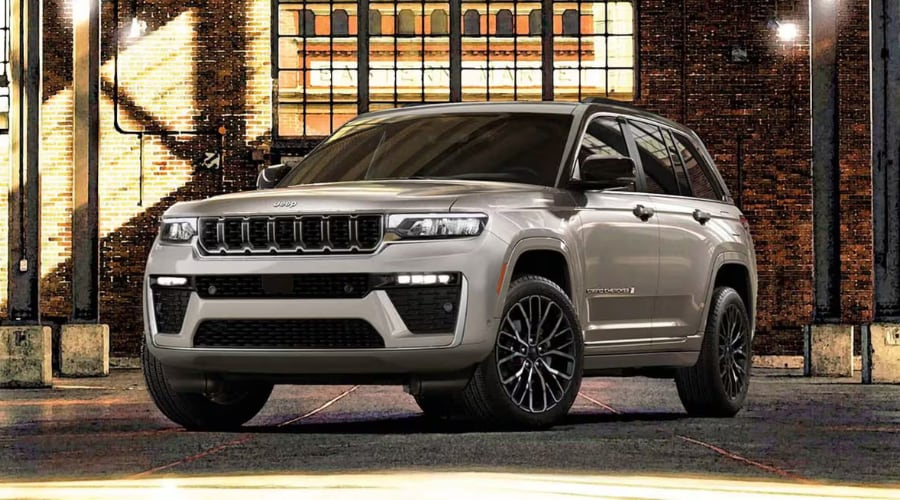 Front angle view of a metallic silver 2026 Jeep Grand Cherokee SUV with dark alloy wheels parked outside a brick building, highlighted for summer maintenance and engine protection in Bergen County, NJ.