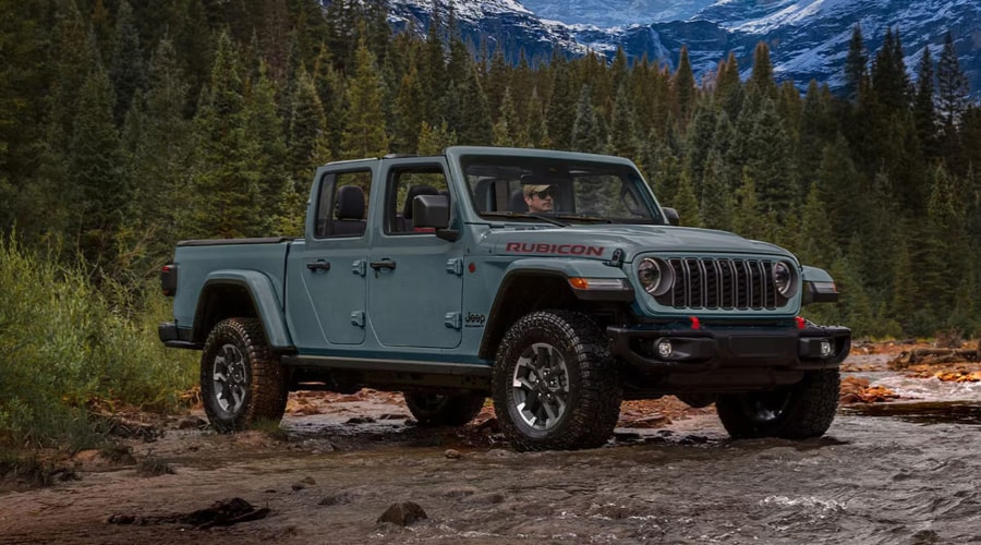 Anvil Gray 2026 Jeep Gladiator Rubicon open-air 4x4 pickup truck crossing a shallow rocky creekbed surrounded by autumn evergreen trees near Ramsey, NJ, showcasing off-road capability, 33-inch all-terrain tires, and red tow hooks.