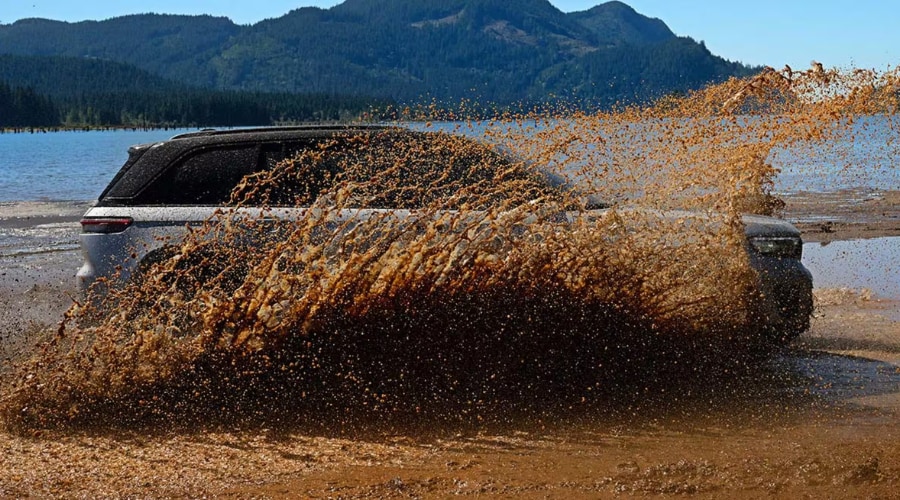 Side profile of a silver 2026 Jeep Grand Cherokee 4x4 splashing through deep muddy water on a shoreline, demonstrating Quadra-Trac traction capability and transfer case fluid protection for drivers in Bergen County, NJ.
