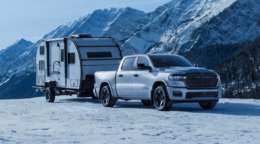 Silver 2026 Ram 1500 towing a camper trailer on a snow-covered road, demonstrating smart towing tech and air suspension control available at Ramsey CJDR in Bergen County, NJ.