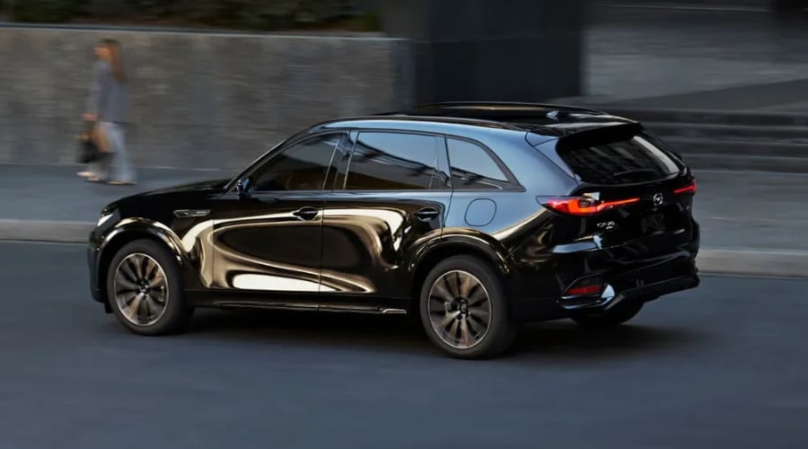 Side profile view of a glossy black 2026 Mazda CX-70 crossover driving smoothly down a street near Ramsey, NJ, demonstrating its responsive handling and executive styling for autumn commuting.