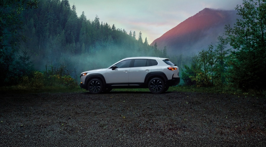 Side profile of a white 2026 Mazda CX-50 Hybrid parked on a wet gravel trail surrounded by misty pine trees near Mahwah, New Jersey. 