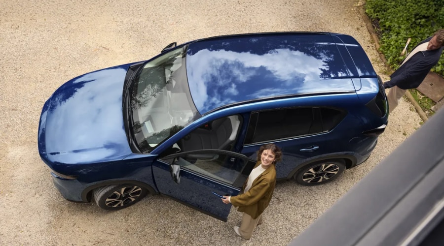 High-angle view of a deep blue 2026 Mazda CX-5 on a gravel driveway in Ramsey, NJ as a driver steps out with a smile before an evening Bergen County commute