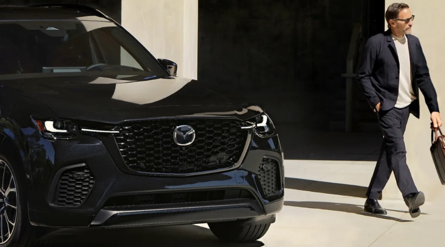 A professional driver carrying a leather briefcase approaches a sleek black 2026 Mazda CX-70 on a bright autumn morning in Ramsey, NJ, preparing for a smooth commute along Route 17.