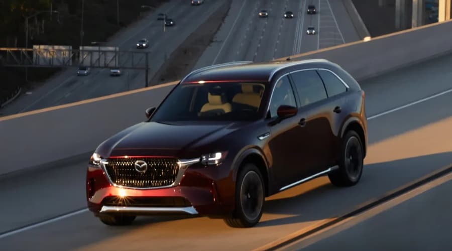 Dark red 2026 Mazda CX-90 three-row SUV with i-Activ AWD driving along an elevated highway overpass near Ramsey, NJ during a late afternoon autumn sunset.